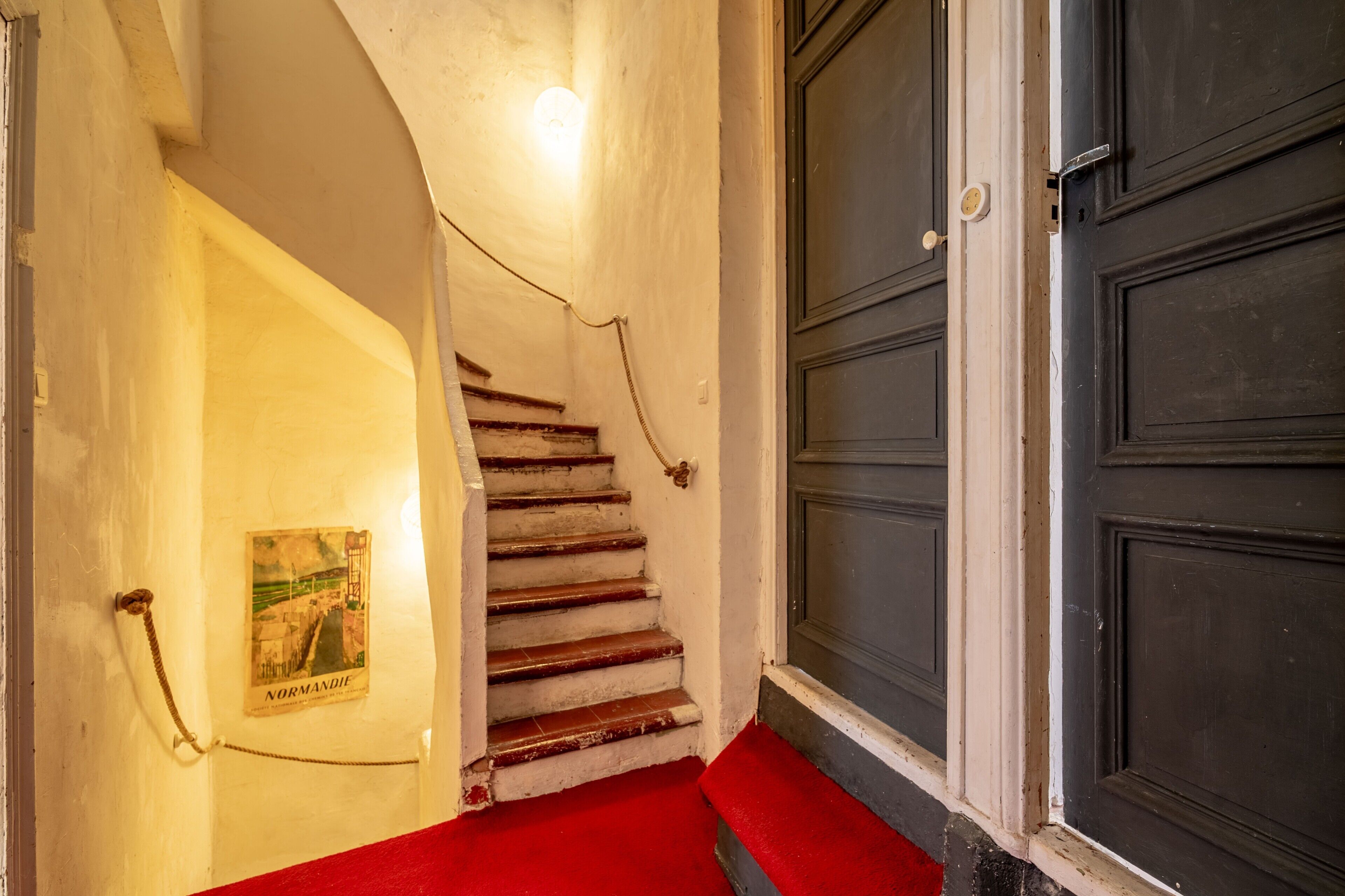 Un escalier menant à une pièce dont la porte est sombre et où se trouve une moquette rouge.