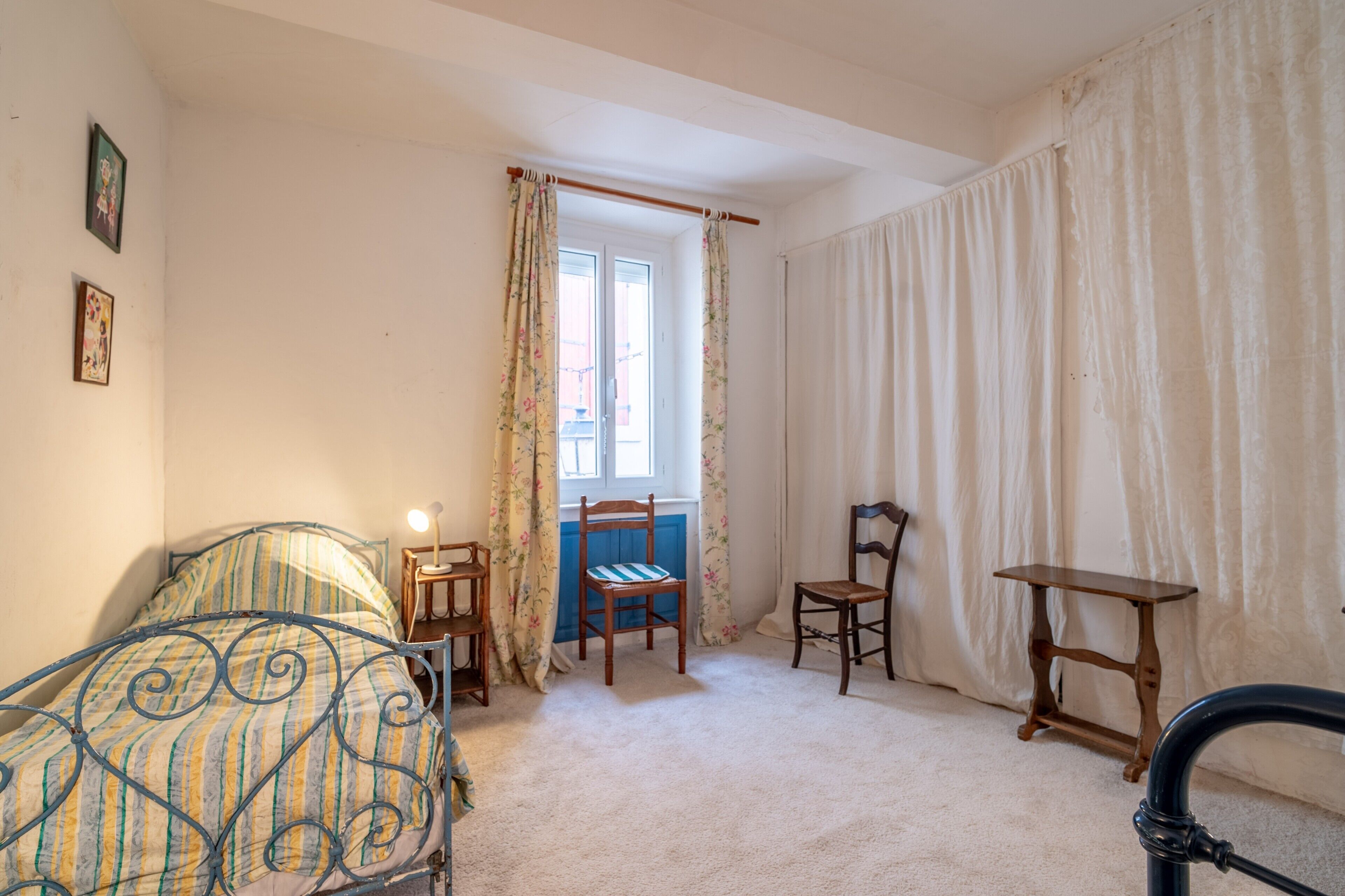 A bedroom with a bed, two chairs, a small table, a window with curtains, and a radiator.