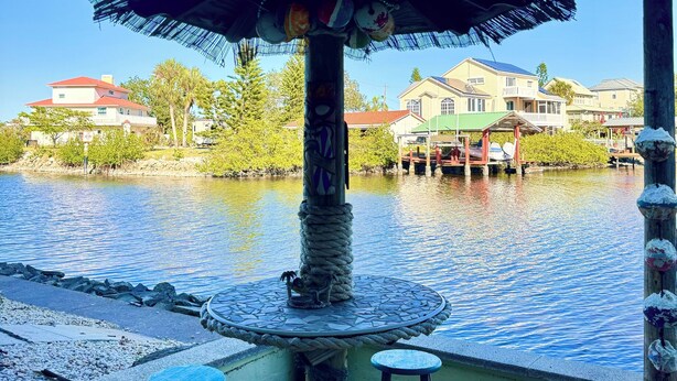 An outdoor seating area with a thatched umbrella, a rope-wrapped column, and a view of a calm body of water and buildings in the background.