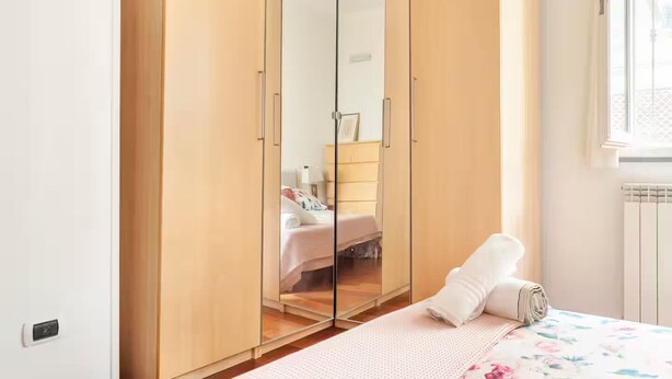 A bedroom with a wooden wardrobe, a bed with a floral bedspread, and a window with a view of the outside.