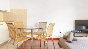 A modern, minimalistic room with a glass dining table surrounded by wicker chairs, a flat-screen TV, and a bed with a quilted cover.
