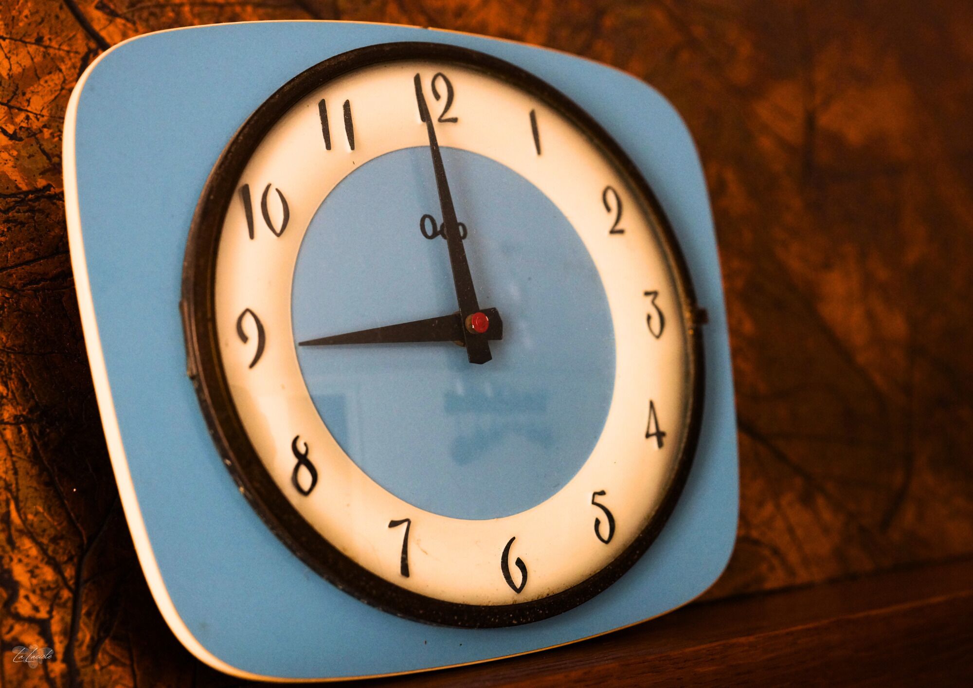 Un reloj de pared cuadrado con esfera azul y blanca, que muestra los números del 1 al 12 y un segundero.