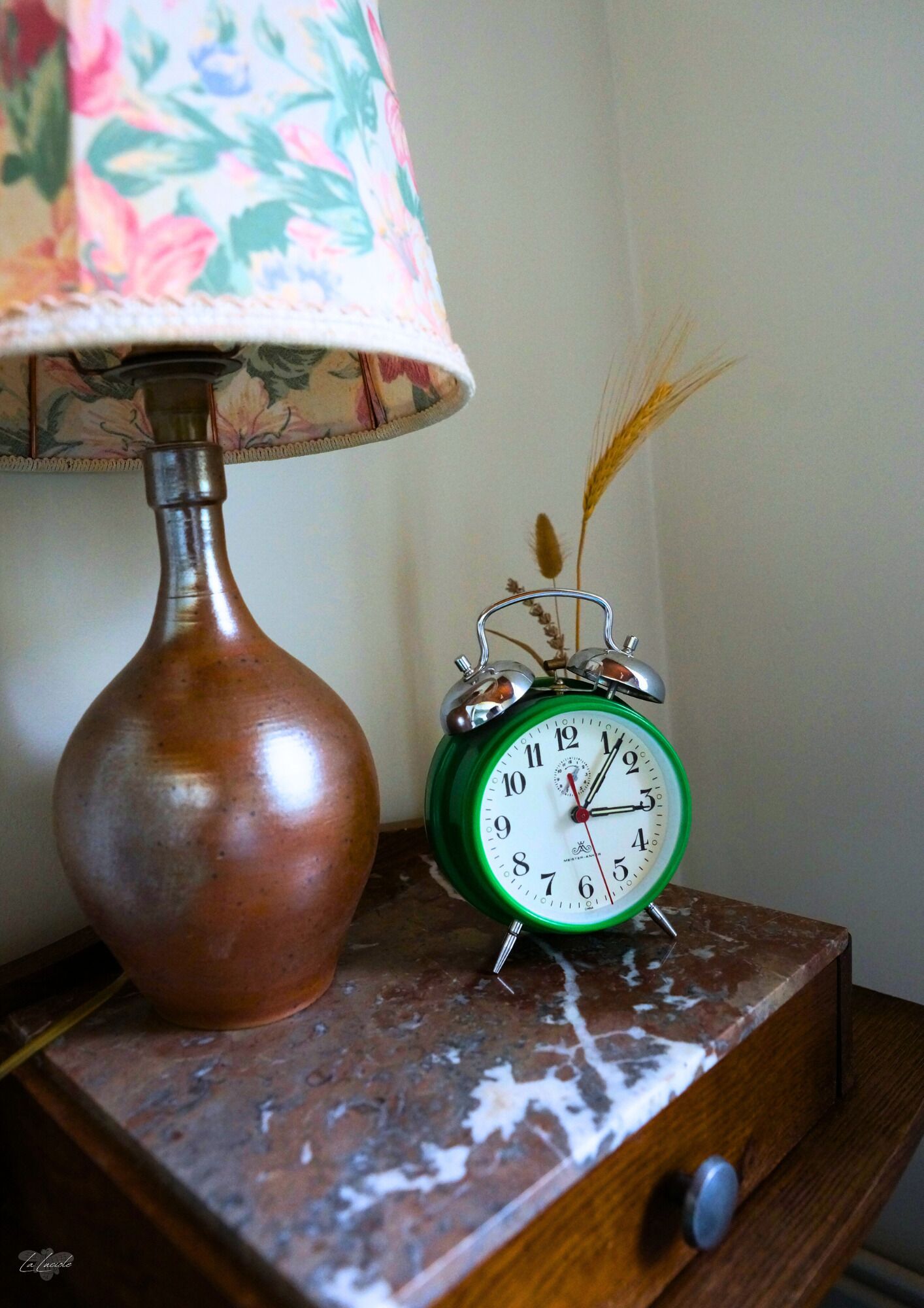 A vintage green alarm clock on a wooden nightstand with a floral lampshade and a copper vase.