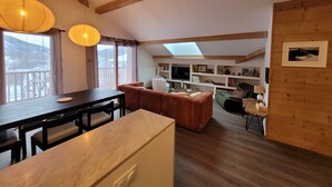 A cozy living room with a dining table, sofas, and a TV. There is a bookshelf and a window with curtains. - Beautiful new T3 1km from the slopes (Le Monêtier-les-Bains)