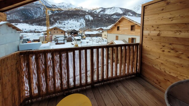 A wooden deck with a view of a snowy mountain village and a construction site. - Beautiful new T3 1km from the slopes (Le Monêtier-les-Bains)