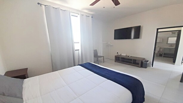 A bedroom with a bed, a TV mounted on the wall, a bench, and a door leading to a bathroom. - Condo Casamar 3 by Kivoya (Puerto Peñasco)