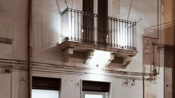 Interieur van een gebouw met een balkon, metalen hekwerk en een lamp erboven. Twee deuren zijn zichtbaar, waarvan er één '15' heeft staan en de andere een gedeeltelijk zichtbaar nummer.