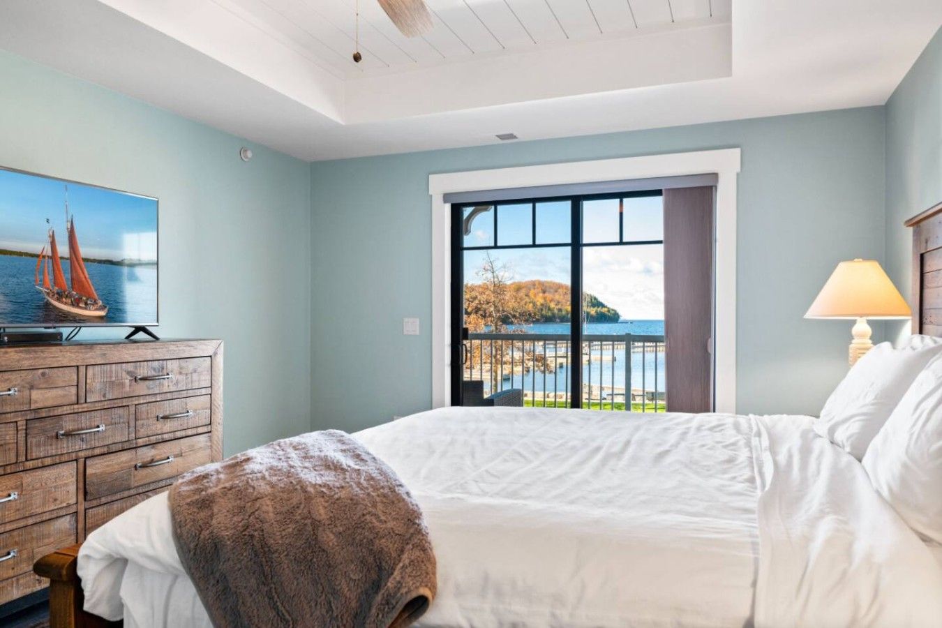 A bedroom with a bed, a dresser, a television, and a window with a view of a body of water.