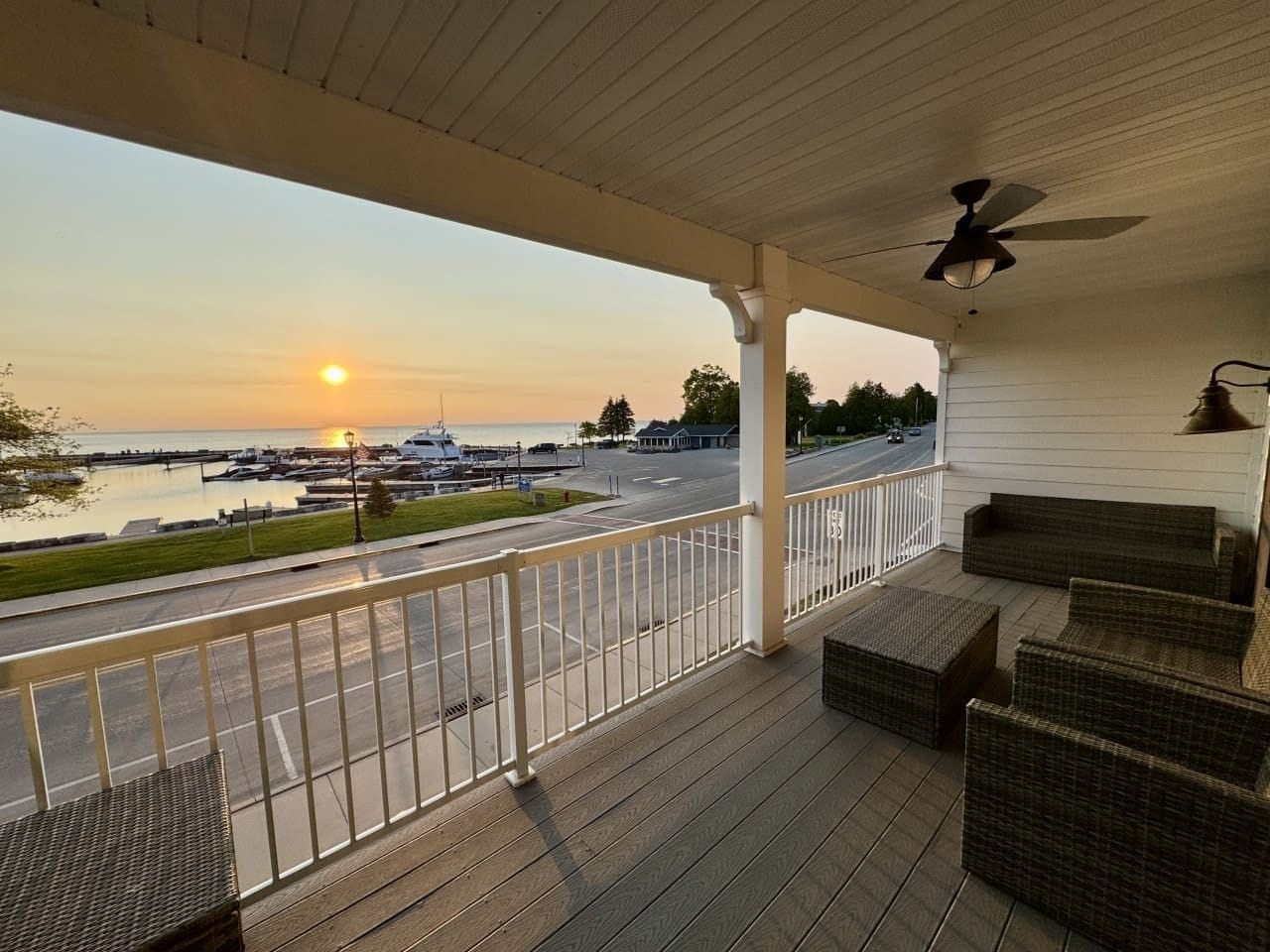 Un porche techado con muebles de mimbre, vista a un puerto deportivo con barcos y una puesta de sol.