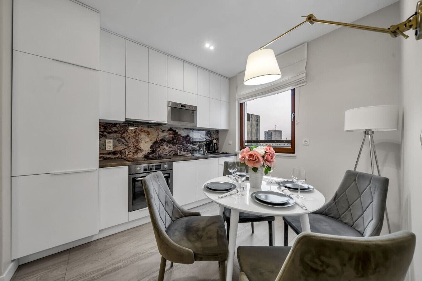 Una cocina moderna con armarios blancos, salpicadero de mármol y un comedor con mesa redonda y sillas tapizadas en gris.