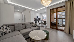 A modern living room with a grey sectional sofa, a round table, and a chandelier. There is a dining area with chairs and a kitchen in the background. - Wola Apartment - 9th Floor, Parking & Balcony - by Rentujemy (Warszawa)