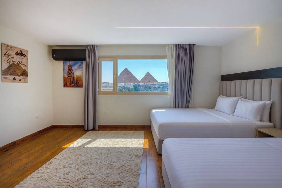 A hotel room with two beds, a large window with a view of pyramids, a wall with posters, and a modern light fixture.