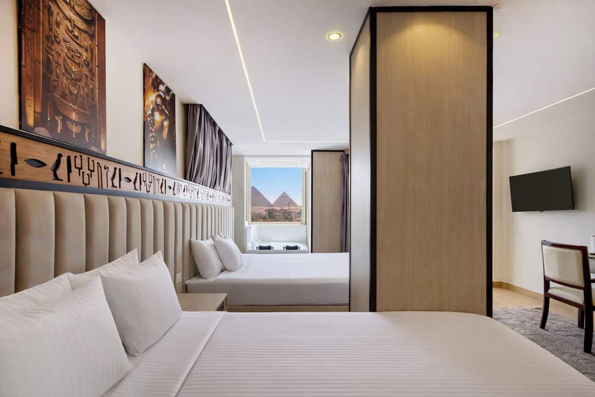 A hotel room with a large bed, a smaller bed, a TV, and a view of pyramids.