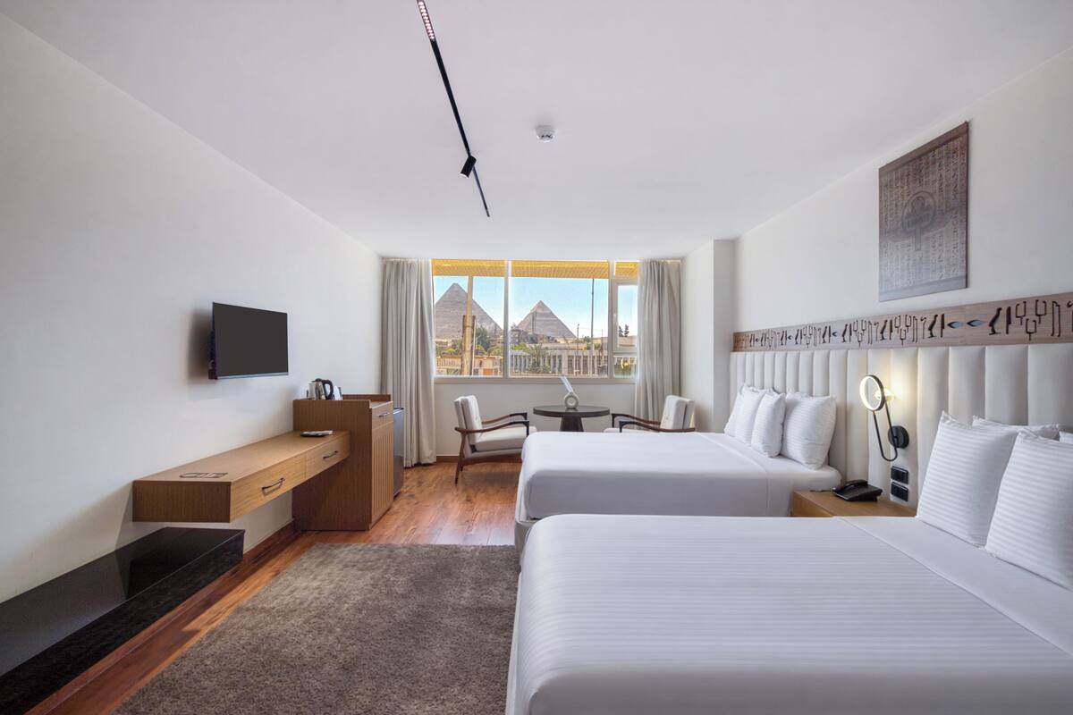 A hotel room with two beds, a desk, a chair, a TV, and a view of pyramids.