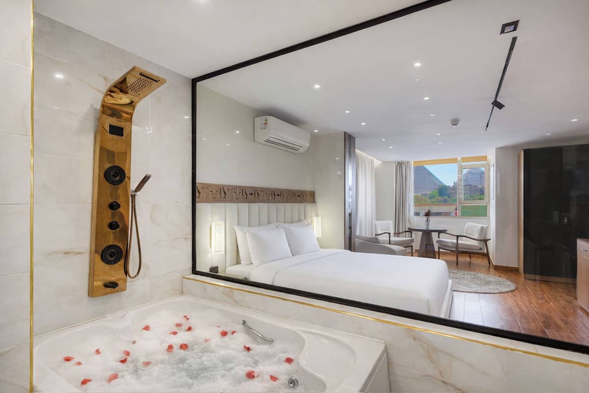 A modern hotel room with a large mirror, a bathtub with rose petals, a wooden shower panel, a bed with white linens, and a view of the outdoors through a window.