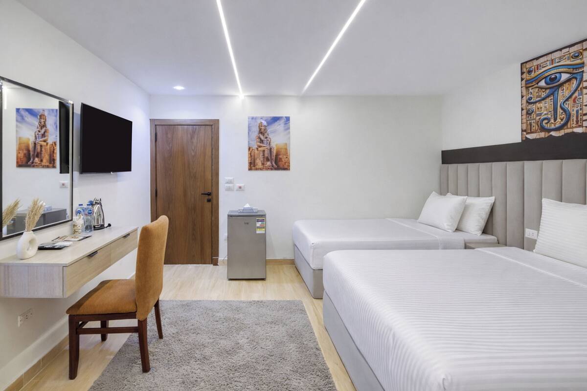A hotel room with a desk, chair, two beds, a TV, artwork, and a refrigerator.