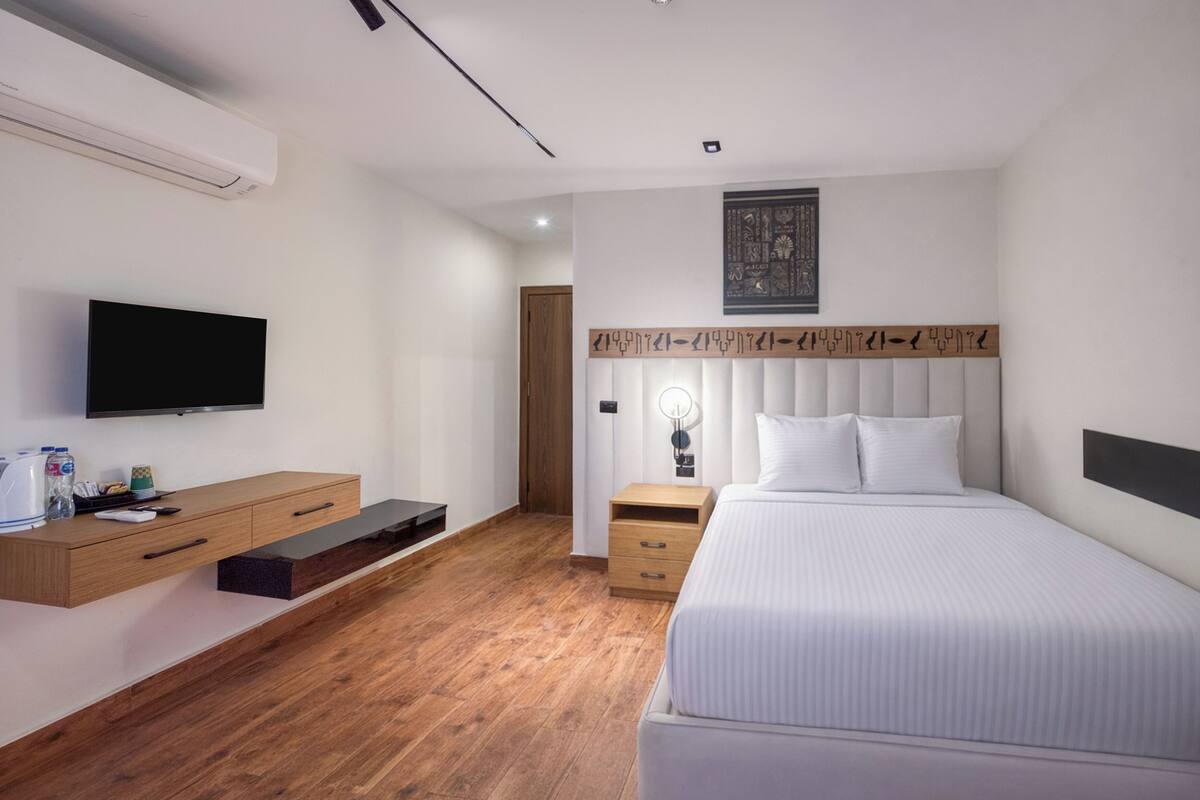 A modern hotel room with a large bed, a flat-screen TV, a wooden dresser, and a bedside table with a lamp.