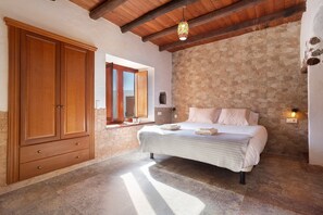 A bedroom with a stone wall, wooden wardrobe, bed with pillows, and a hanging light fixture.