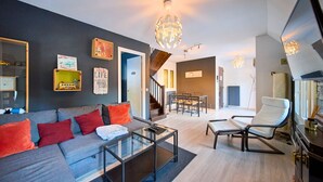 Una sala de estar moderna con un sofá gris, cojines rojos, una mesa de centro de cristal y un sillón blanco. Se ve una escalera que lleva a otra habitación, un comedor con mesa y sillas, y un espejo grande en la pared.