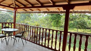 Un espacio al aire libre techado con piso de madera, una mesa redonda con sillas, y vista a un jardín con árboles.