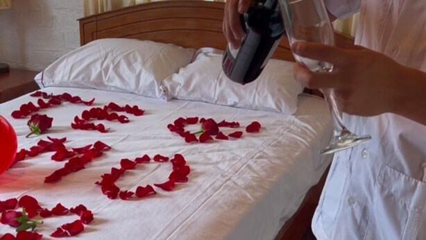 Una persona sirve una bebida en un vaso sobre una cama con pétalos de rosa dispuestos formando la palabra 'LOVE'.