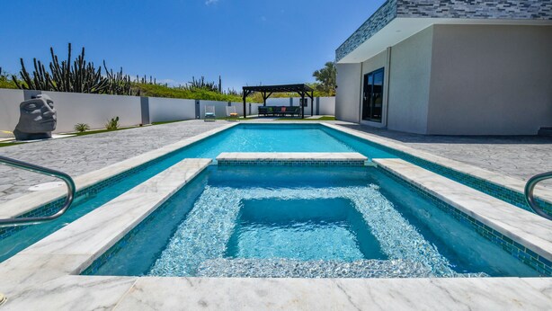 Una piscina rectangular con borde de azulejos, rodeada por un muro bajo y una pérgola con asientos.