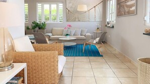 A spacious living area with a sofa, two wicker chairs, a round coffee table, and a blue rug. There are two windows with white shutters, a hanging lamp, and a painting on the wall.