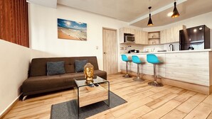 A modern living room with a sofa, a coffee table, and a Buddha statue. There are three blue bar stools and a kitchen area with a refrigerator and microwave. - Departamento 5ta Av-Playa & Rooftop Pool (Playa del Carmen)