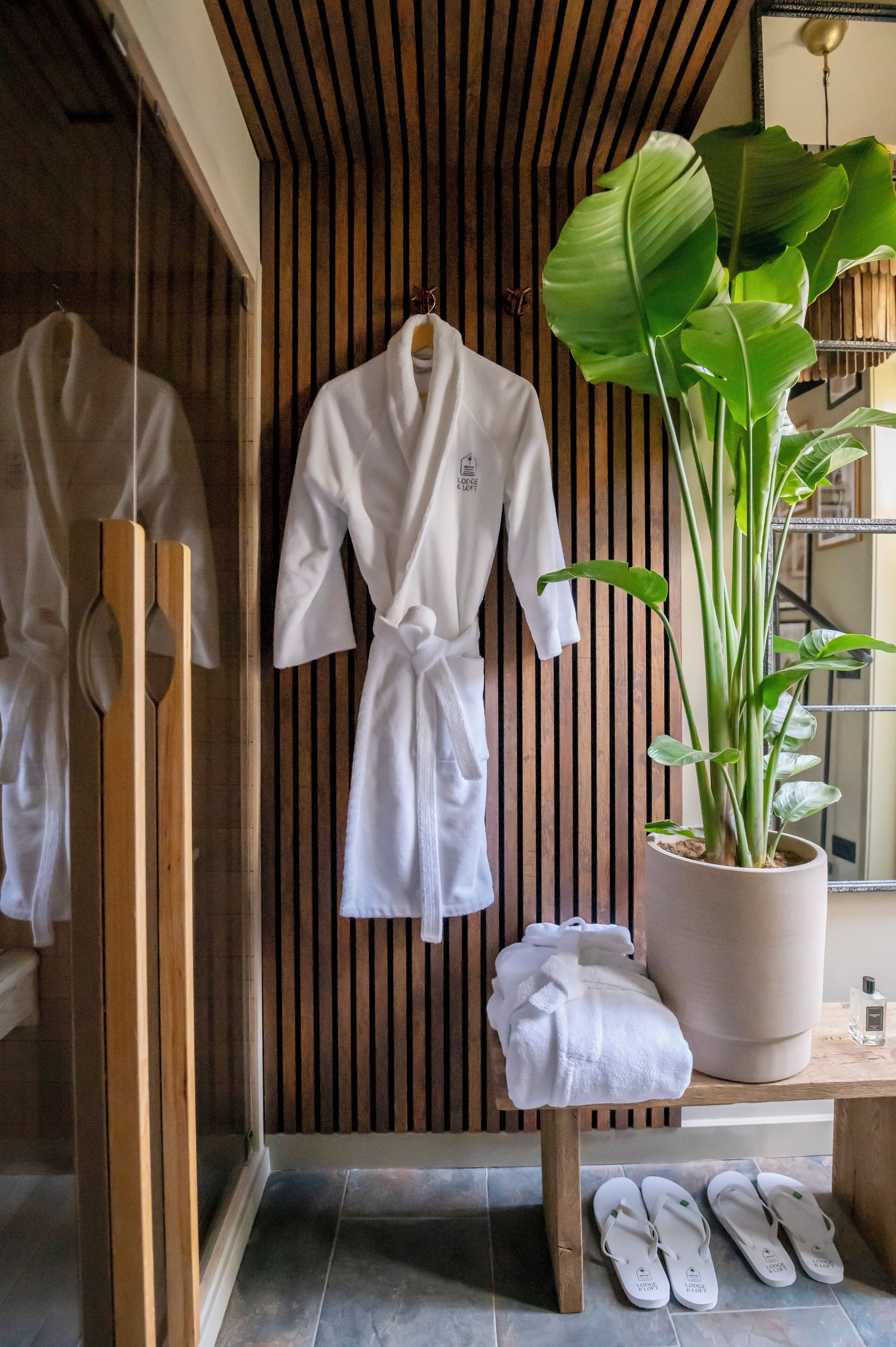 Ein Badezimmer mit Holzvertäfelung, einem Topf mit Pflanze, einem Bademantel auf einem Gestell, einem Handtuch auf einer Bank und einem Paar Hausschuhen auf dem Boden.