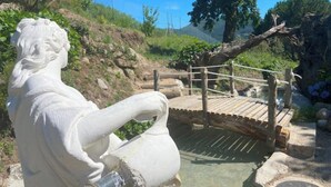 Eine Skulptur eines Mannes, der eine Kanne hält, eine Holzbrücke und eine natürliche Landschaft mit Bäumen und einem Hügel.