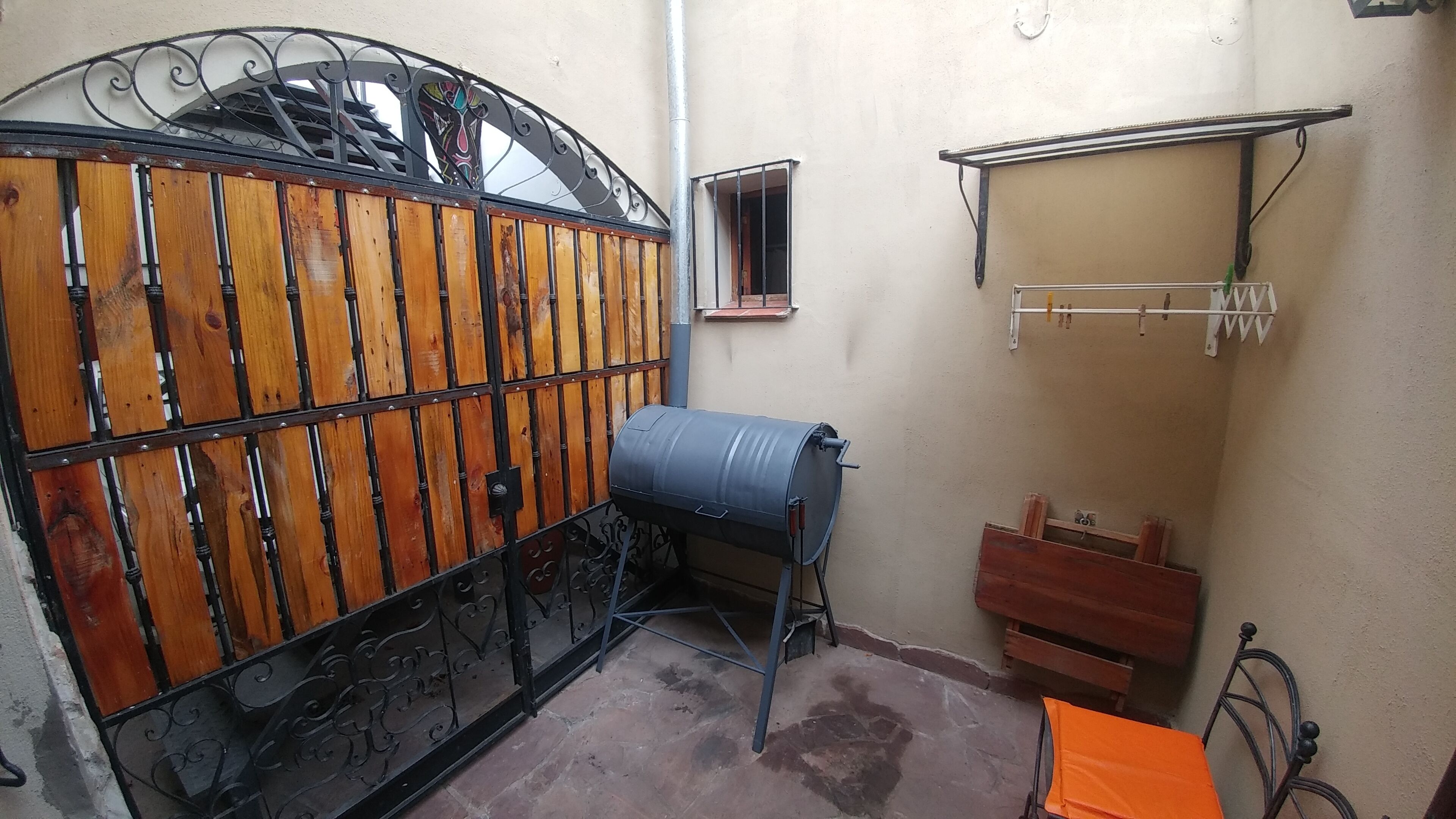 Uma pequena área externa fechada, com portão de madeira, barril de metal, suporte de metal e banco de madeira.