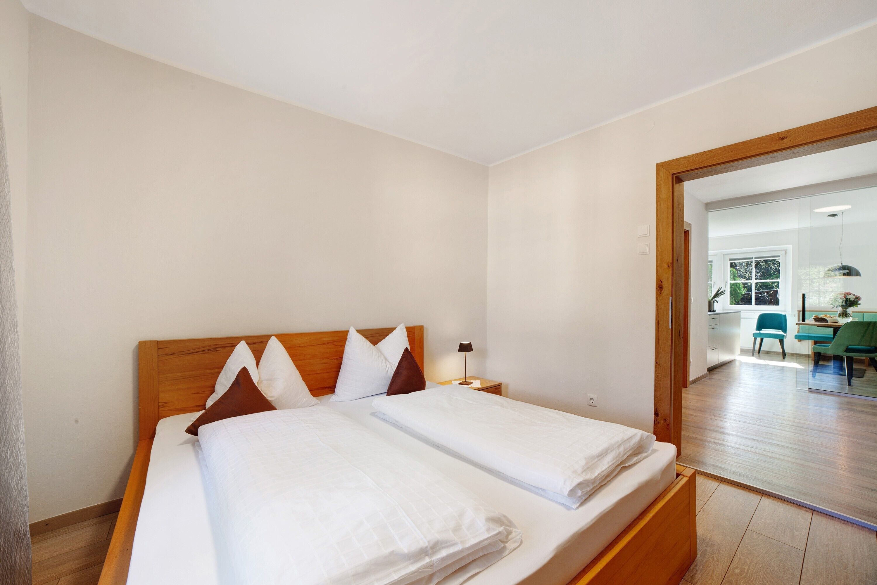 A double bed with a wooden headboard, a nightstand with a lamp, and a view of a dining area with a chair and table.