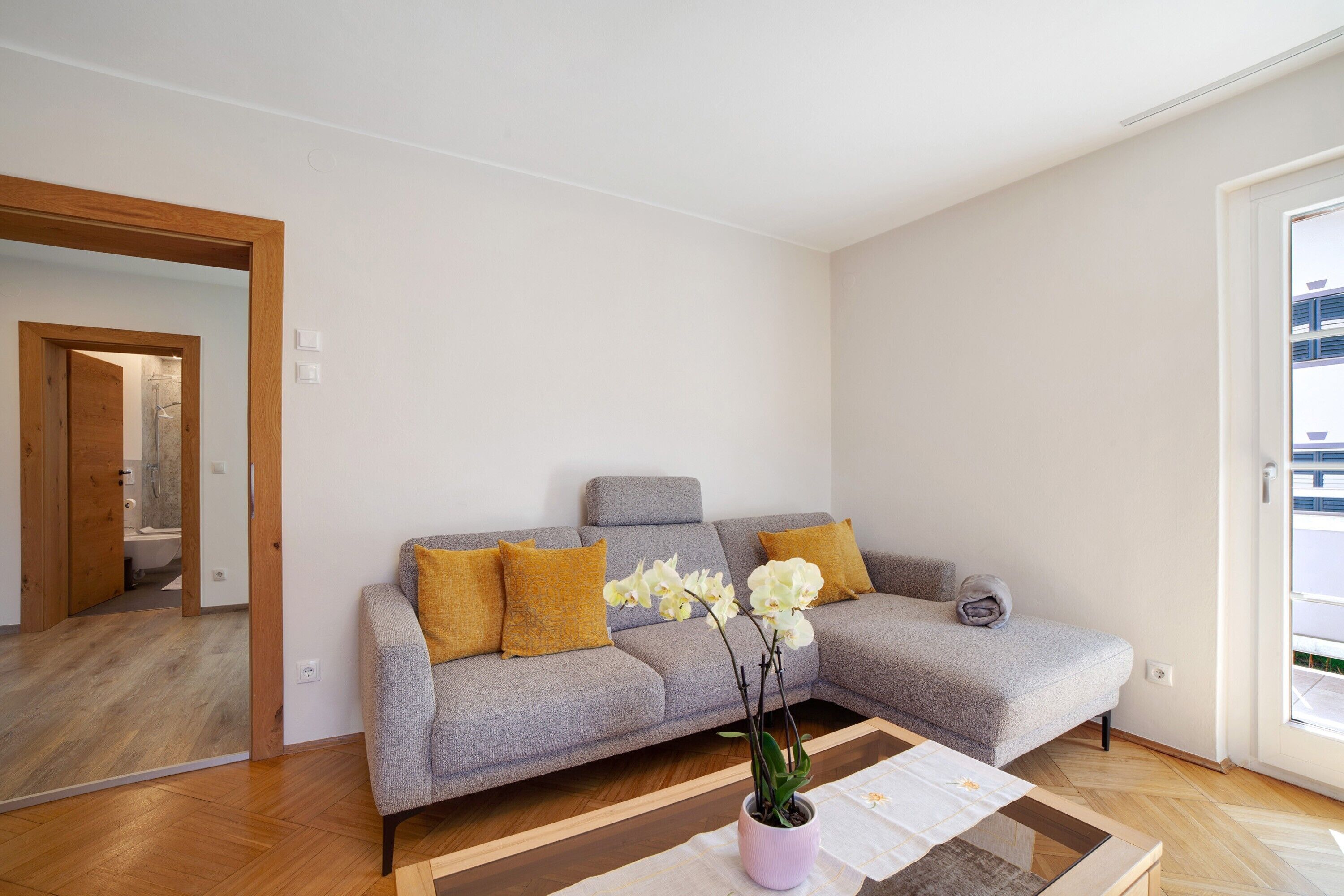 Una sala de estar moderna con un sofá gris, piso de madera y una orquídea en maceta.