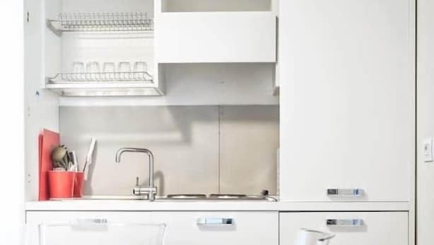 A modern kitchen with white cabinets, a stainless steel sink, and a countertop. There are clear plastic chairs and a dining table.