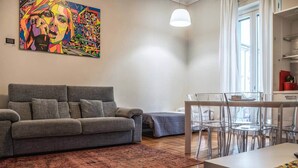 A modern living room with a grey sofa, a dining table with clear chairs, a colorful abstract painting on the wall, and a large window with curtains.