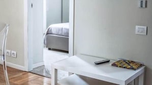 A room with a white bench, a flat-screen TV mounted on the wall, a bed with a grey mattress, and a door leading to another room.