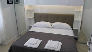 A neatly made bed with a grey bedspread, two white pillows, and folded white towels on the foot of the bed. There are two bedside tables with lamps, and a white wall with a light switch and a small shelf.