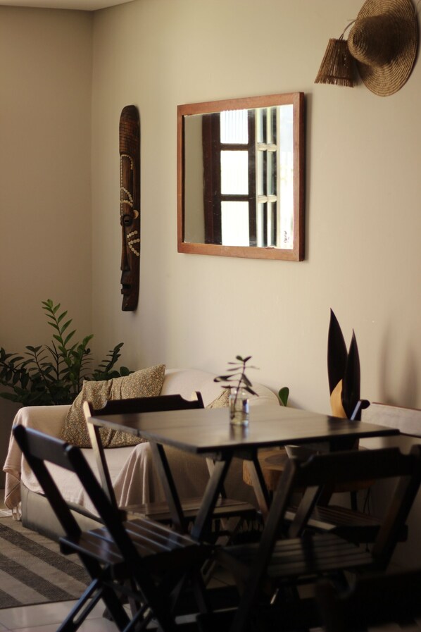 Una habitación con una cama, una mesa de comedor, sillas, un espejo, una escultura de madera en la pared y un sombrero colgado.