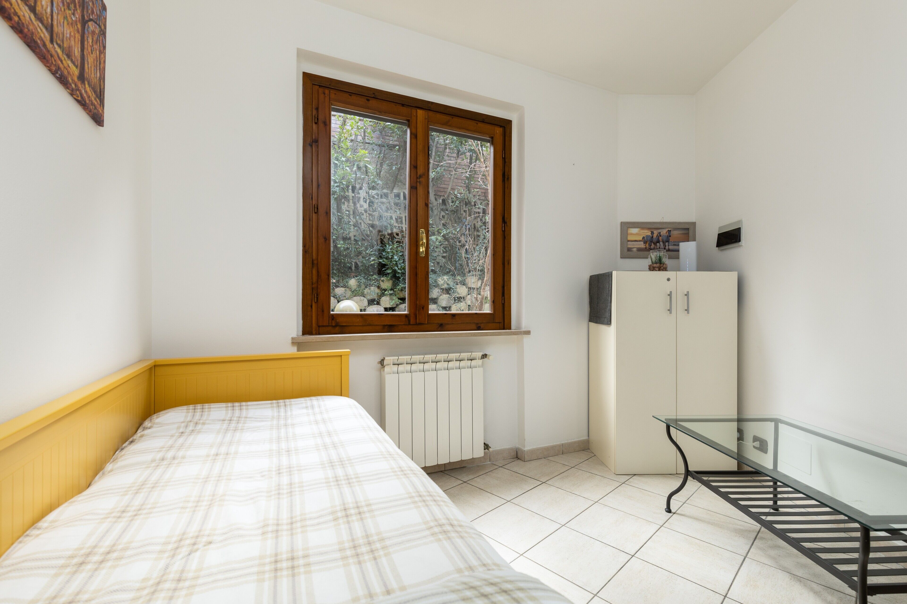 Ein Schlafzimmer mit Bett, Heizkörper, Fenster mit Blick ins Grüne, Schrank und Glastisch.
