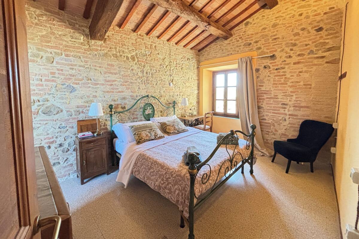 A bedroom with a stone wall, a wooden ceiling, a bed with a patterned bedspread, a nightstand, a chair, and a window with curtains.