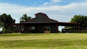 A barn with a red roof and a covered area, surrounded by a grassy field. - Peaceful Texas Retreat w/Gourmet Kitchen, Ranch Views, Optional MasterChef Meals (Lipan)