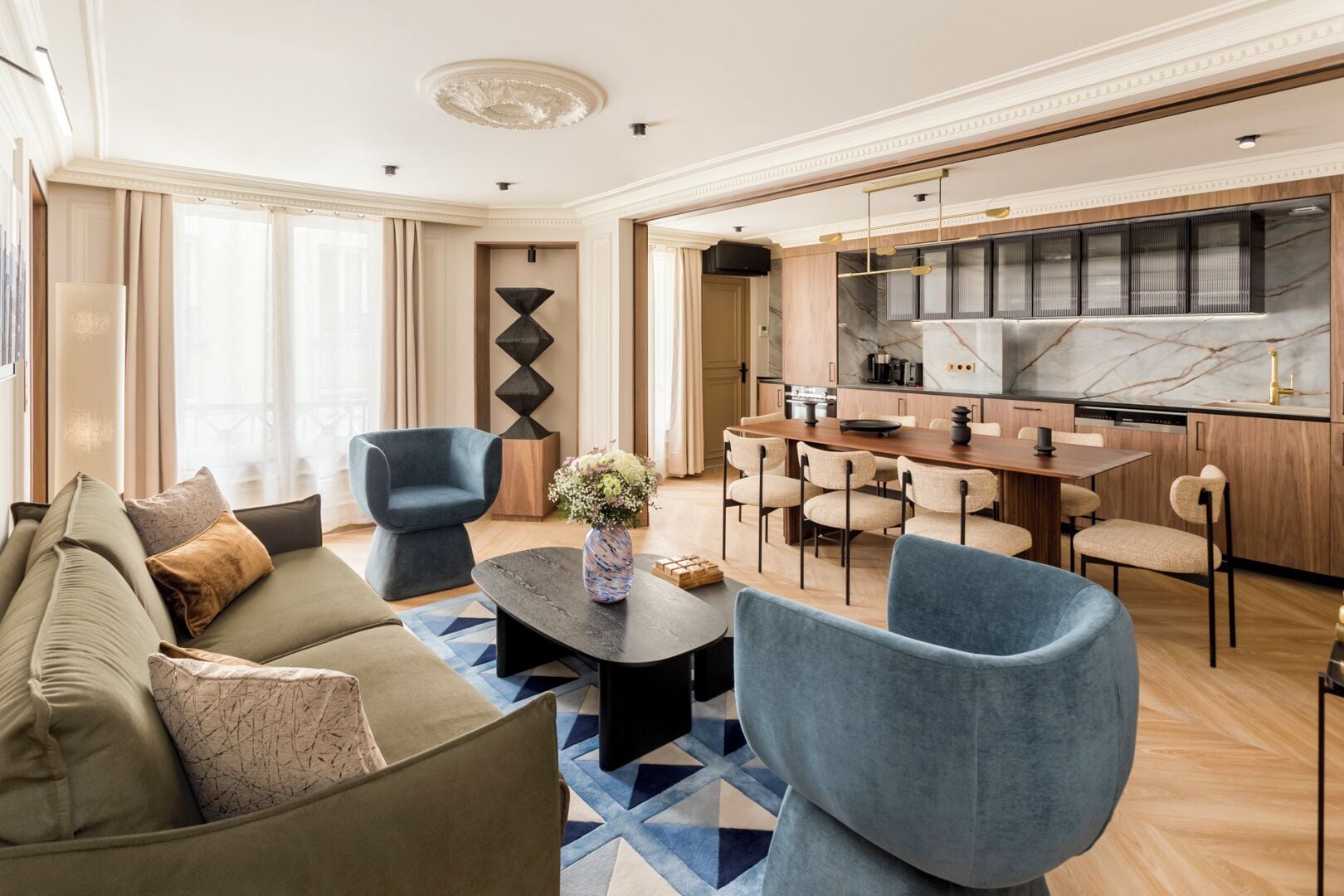 Una sala de estar moderna con un sofá, sillas y una mesa de centro. Se observa una zona de cocina con armarios de madera y una encimera de mármol.