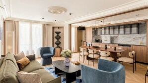 A modern living room with a sofa, chairs, and a coffee table. There is a kitchen area with wooden cabinets and a marble countertop. - Merveil - Signature Suite - Opera - Bateliere (Paris)