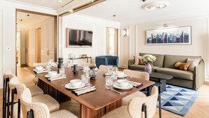 A spacious dining area with a long wooden table set for a meal, flanked by upholstered chairs. A television is mounted on the wall above the table, and a sofa with cushions is positioned in the background. - Merveil - Signature Suite - Opera - Bateliere (Paris)