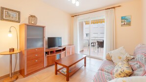 A living room with a sofa, a wooden coffee table, a TV on a cabinet, a lamp, and a balcony with a table and chairs.