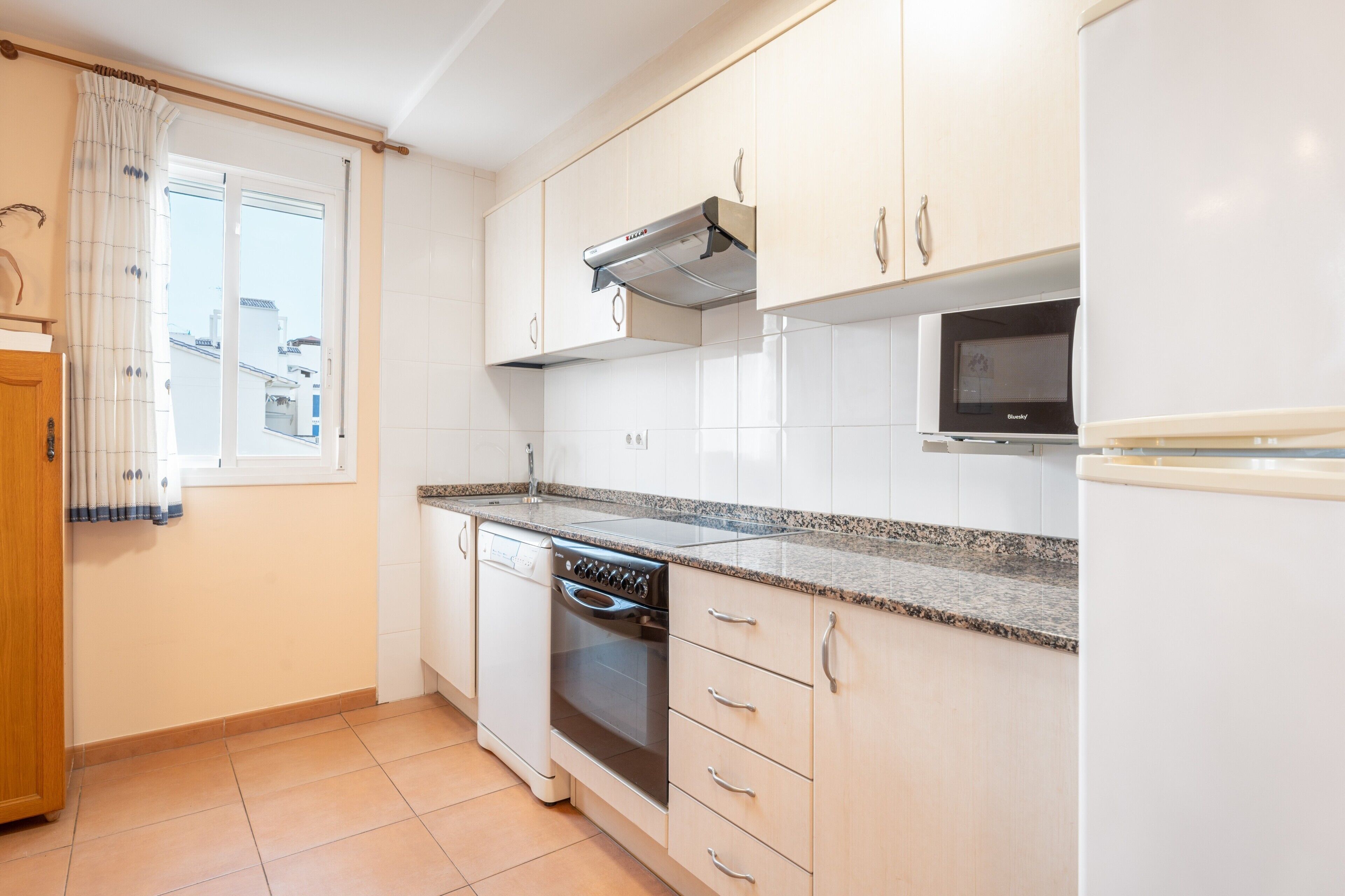 Una cocina con armarios blancos, lavavajillas, horno, microondas y una ventana con cortinas.