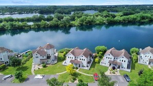 A residential area with houses by a lake, featuring a central house with a large porch and a garage. - Lake Access, Hot Tub, 4+MBed, 3Bth, Landings, Cotswolds Water Park, Sth Cerney (South Cerney)