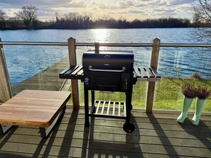 A barbecue grill on a wooden deck with a view of a lake. - Lake Access, Hot Tub, 4+MBed, 3Bth, Landings, Cotswolds Water Park, Sth Cerney (South Cerney)