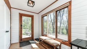 Una habitación con pisos de madera, un banco, una puerta y ventanas grandes que dan a vista de árboles.