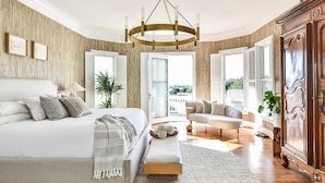 Ein Schlafzimmer mit einem großen Bett, Nachttischen, einer Bank, einer Chandelier und Blick durch die geöffnete Tür.